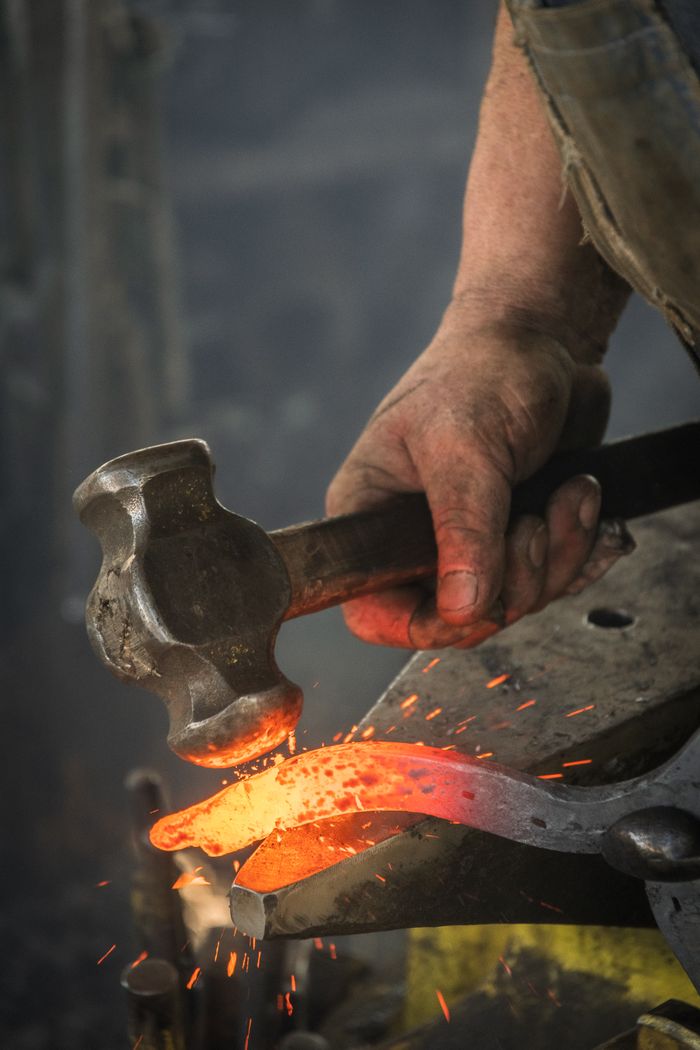 our-mission-01 Close-up of a blacksmith hammering a hot iron piece in a workshop.